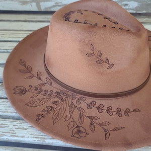 May include: A brown fedora-style hat with a decorative floral design etched into the brim and crown. The hat features a brown leather band and is made of a soft, suede-like material. The hat is a stylish accessory.