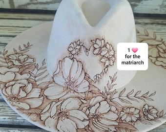 May include: A cream-colored hat with a wide brim, featuring intricate floral designs in shades of brown. The hat has a heart-shaped floral design and the text "1 for the matriarch".