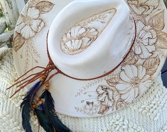 May include: A white felt cowboy hat with brown leather trim and floral designs burned into the felt. The hat has a brown leather strap with a beaded tassel and a long feather accent.