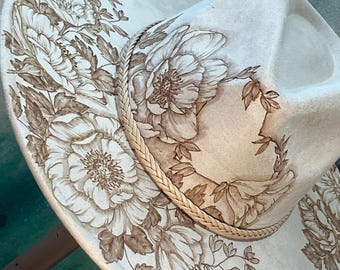May include: A cream-colored wide-brim hat featuring intricate brown floral illustrations covering the crown and brim. A braided band encircles the base of the crown, separating the top from the brim. The floral design includes detailed petals and leaves.