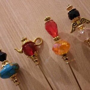 May include: Four decorative pins with gold metal bases and colorful glass beads. The beads are shaped like a teapot, a teardrop, a round bead, and an angel with wings.