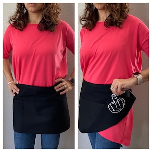 May include: A pink short-sleeved t-shirt with a black apron featuring a white middle finger graphic. The apron has a pocket on the front.
