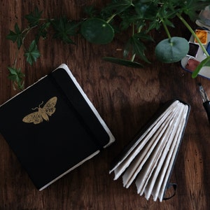 May include: A black notebook with a gold moth illustration on the cover, a second open notebook with visible white pages, a metal watercolor paint tin with six colorful pans, and a black and blue paintbrush with a metal ferrule.