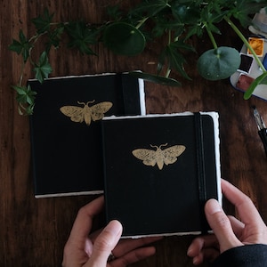 May include: Two square black notebooks with deckled white paper edges and a gold moth illustration on the front cover. Each notebook features a black elastic band closure to keep the pages secure. The covers are matte black with a gold foil moth.