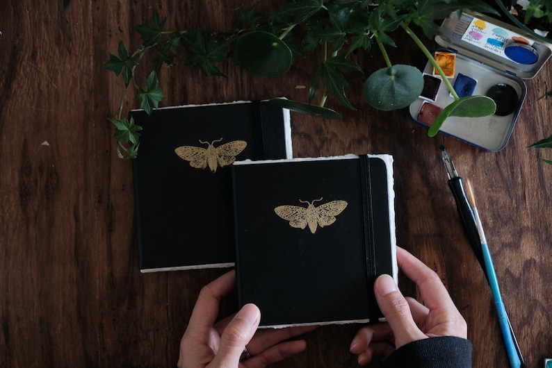 May include: Two square black notebooks with deckled white paper edges and a gold moth illustration on the front cover. Each notebook features a black elastic band closure to keep the pages secure. The covers are matte black with a gold foil moth.