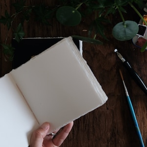 May include: An open sketchbook with thick, off-white pages featuring rough, deckled edges. Beside the book lies a black dip pen, a thin blue-handled paintbrush, and a small metal watercolor paint tin containing various colorful pigment pans.
