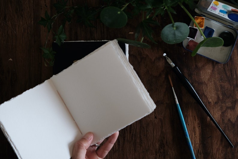 May include: An open sketchbook with thick, off-white pages featuring rough, deckled edges. Beside the book lies a black dip pen, a thin blue-handled paintbrush, and a small metal watercolor paint tin containing various colorful pigment pans.