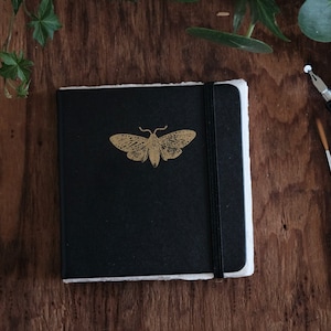 May include: A square black notebook with a gold moth illustration on the front cover. The notebook features a black elastic band closure and thick, textured white paper edges visible on the side. The cover has a smooth, matte finish.