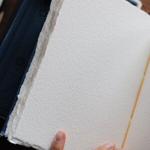 May include: Open handmade paper journal with textured, off-white pages. The pages have a rough, deckled edge. The spine is bound with a yellow thread. A blue book is visible in the background.