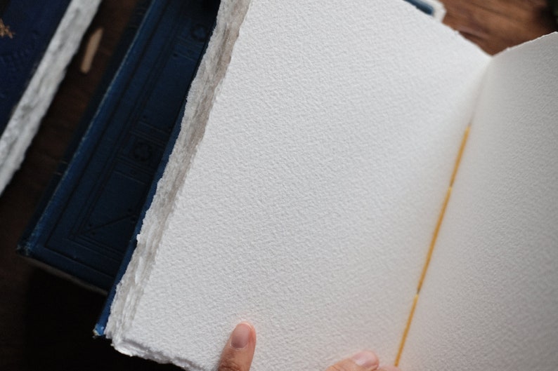 May include: Open handmade paper journal with textured, off-white pages. The pages have a rough, deckled edge. The spine is bound with a yellow thread. A blue book is visible in the background.
