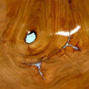 Puede incluir: Primer plano de un cuenco de madera con un patr&oacute;n de veta de madera natural. El cuenco tiene un color marr&oacute;n claro y un acabado liso y pulido. Una peque&ntilde;a incrustaci&oacute;n de resina azul claro es visible en el centro del cuenco.