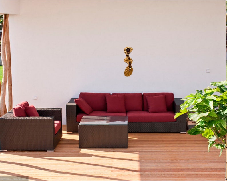 May include: A red wicker sofa with three cushions and a matching wicker armchair with a cushion. The furniture is on a wooden deck with a gold metal wall sculpture hanging on the white wall behind the sofa.