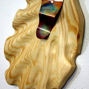 May include: A wooden wall sculpture with a wavy, organic shape. A piece of iridescent metal is attached to the wood, creating a contrasting texture.