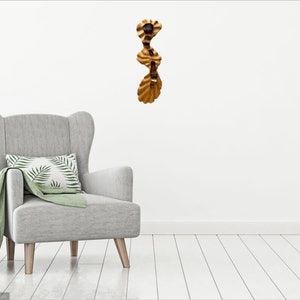 May include: A gold-colored metal wall sculpture with a wavy, organic design. The sculpture is made of multiple pieces that are connected together. It is hanging on a white wall in a room with a gray armchair.