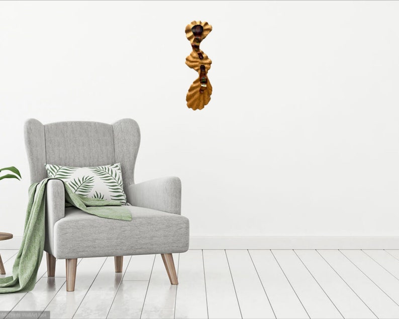 May include: A gold-colored metal wall sculpture with a wavy, organic design. The sculpture is made of multiple pieces that are connected together. It is hanging on a white wall in a room with a gray armchair.