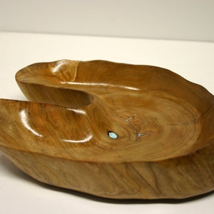 Puede incluir: Un cuenco de madera con una forma &uacute;nica y asim&eacute;trica y un color marr&oacute;n claro. El cuenco tiene un acabado liso y pulido y una peque&ntilde;a incrustaci&oacute;n turquesa.