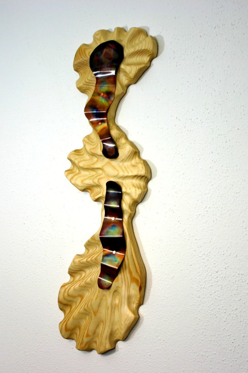 May include: A wall sculpture made of wood and metal. The wood is a light brown color and has a wavy texture. The metal is a copper color and has a shiny finish. The metal is shaped into three wavy lines that are attached to the wood.