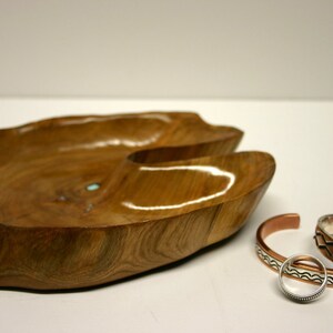 Puede incluir: Un cuenco de madera con una forma natural y org&aacute;nica, de color marr&oacute;n claro. El cuenco tiene una peque&ntilde;a incrustaci&oacute;n turquesa. Tres pulseras de plata y cobre se colocan junto al cuenco.