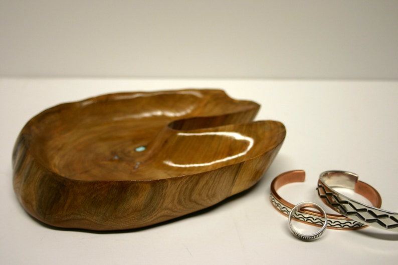 Puede incluir: Un cuenco de madera con una forma natural y org&aacute;nica, de color marr&oacute;n claro. El cuenco tiene una peque&ntilde;a incrustaci&oacute;n turquesa. Tres pulseras de plata y cobre se colocan junto al cuenco.