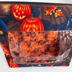May include: A black zippered pouch with a clear plastic window. The pouch is decorated with a Halloween theme featuring orange pumpkins with jack-o-lantern faces and a background of blue, brown, and orange.
