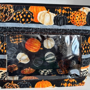 May include: Black zippered pouch with a clear plastic window. The pouch is decorated with a repeating pattern of orange, white, and black pumpkins.