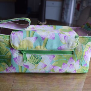 May include: A rectangular fabric storage caddy featuring a light green and pink lotus flower pattern. The caddy includes a front zippered pocket with a green zipper pull, a top flap closure, and a side handle with a snap button fastener.