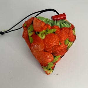 May include: A small, diamond-shaped drawstring bag with a vibrant strawberry pattern. The bag features a black drawstring and a red plastic toggle. The fabric showcases detailed illustrations of ripe, red strawberries with green leaves.