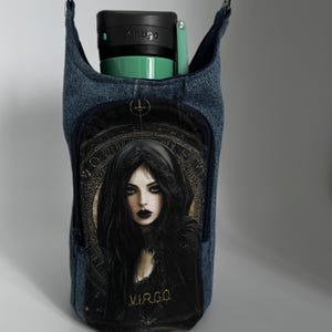May include: A denim blue crossbody bag with a zipper closure. The bag features a black and gold print of a woman with the word "VIRGO" underneath. The bag is designed to hold a water bottle.