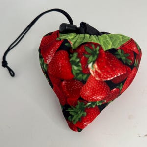 May include: A strawberry-shaped drawstring bag with a black drawstring and stopper. The bag is covered in a pattern of red strawberries with green leaves on a black background. The bag is made of fabric.