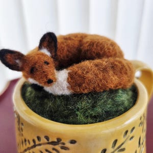 May include: A needle-felted fox with rust-orange and white fur curled up on a textured dark green base. The fox is resting inside a yellow ceramic mug that features a dark brown leaf pattern around the exterior surface of the vessel.