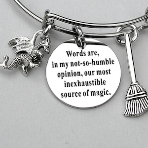 May include: Silver-toned bangle bracelet with three charms. One charm is a dragon, another is a broom, and the third is a round disc with the quote: "Words are, in my not-so-humble opinion, our most inexhaustible source of magic."