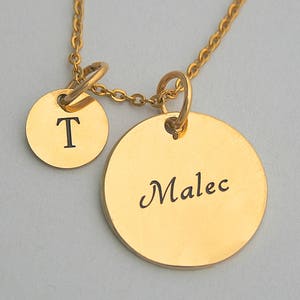 May include: Gold-colored necklace with two circular pendants. One pendant has the letter "T" engraved, and the other pendant has the name "Malec" engraved. The necklace chain and pendants are gold-toned.