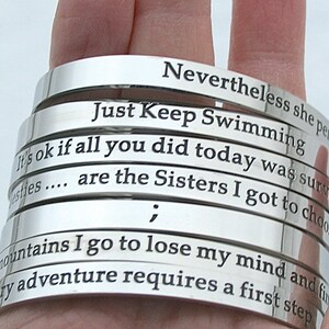 May include: A stack of silver bracelets with engraved inspirational quotes, including "Just Keep Swimming", "It's ok if all you did today was survive", and "Every adventure requires a first step".