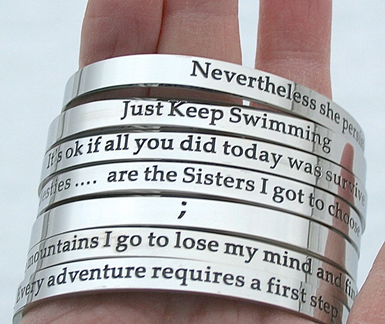 May include: A stack of silver bracelets with engraved inspirational quotes, including "Just Keep Swimming", "It's ok if all you did today was survive", and "Every adventure requires a first step".