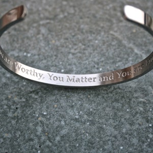 May include: A silver bangle bracelet with the inscription "You are Worthy, You Matter and You are Loved".