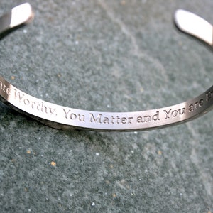 May include: A silver bangle bracelet with the inscription "You are Worthy, You Matter and You are Fine".