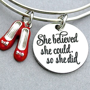 May include: Silver bangle bracelet with two small red shoe charms and a round charm. The round charm has the text "She believed she could, so she did" in black script.