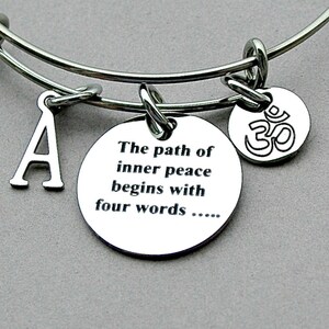 May include: Silver-toned bangle bracelet with three charms. One charm is a capital letter "A", another is a round charm with the Om symbol, and the third is a round charm with the text "The path of inner peace begins with four words....."