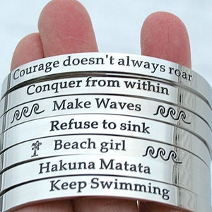May include: A stack of silver bracelets with inspirational quotes engraved on them. The quotes include "Courage doesn't always roar", "Conquer from within", "Make Waves", "Refuse to sink", "Beach girl", "Hakuna Matata", and "Keep Swimming".