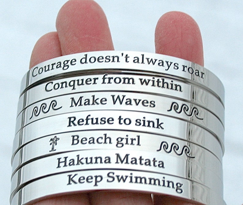 May include: A stack of silver bracelets with inspirational quotes engraved on them. The quotes include "Courage doesn't always roar", "Conquer from within", "Make Waves", "Refuse to sink", "Beach girl", "Hakuna Matata", and "Keep Swimming".