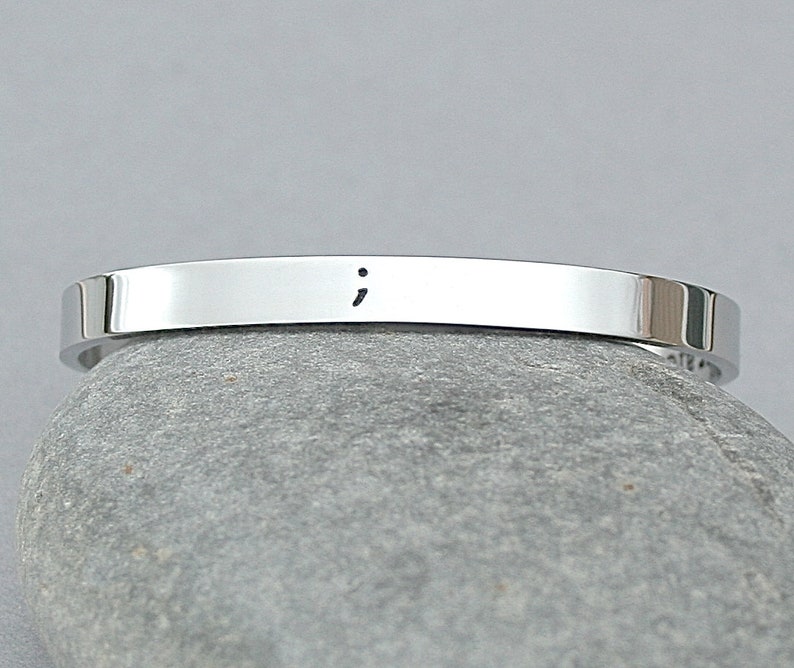 May include: A silver bracelet with a semicolon engraved on it. The bracelet is resting on a gray rock.