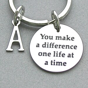 May include: Silver-coloured keyring with a round charm and an initial "A" charm. The round charm is engraved with the words "You make a difference one life at a time".