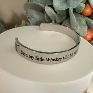 May include: A silver-colored cuff bracelet with the engraved text "She's my little Whiskey Girl. My everything." The bracelet is displayed on a white surface, with greenery and orange fruit in the background.