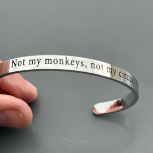 May include: A silver-colored cuff bracelet with the engraved phrase "Not my monkeys, not my circus." The bracelet is curved and open-ended, designed to be slipped onto the wrist. The text is in a simple, sans-serif font.