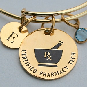May include: Gold charm bracelet with a gold charm that says "Certified Pharmacy Tech" with a mortar and pestle and an "Rx" symbol. The bracelet also has a gold charm with the letter "E".