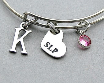 May include: A silver-toned bangle bracelet with three charms. One charm is the letter "K", another is a heart with "SLP" engraved, and the third is a pink gemstone. The bracelet is on a gray background.