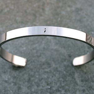 May include: A silver bangle bracelet with a semicolon engraved on the front.