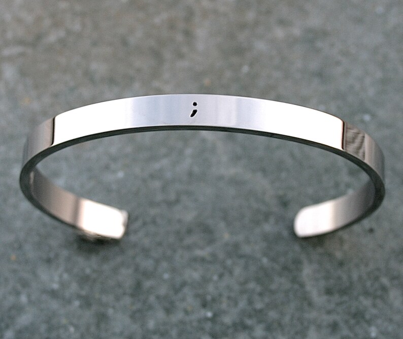 May include: A silver bangle bracelet with a semicolon engraved on the front.