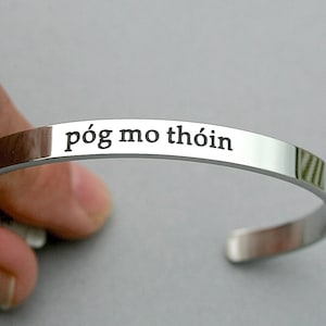 May include: A silver bangle bracelet with the inscription "póg mo thóin" in black lettering.