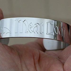 Peut inclure: Bracelet en argent gravé avec les mots "Meat Loaf" en police gothique. Une silhouette de chauve-souris est également gravée sur le bracelet.